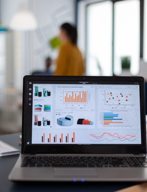 Laptop with marketing charts, graphs analitics in start up company. Workplace area in business center with multi ethnic employees in it, shot of room with modern furniture and blue wall.
