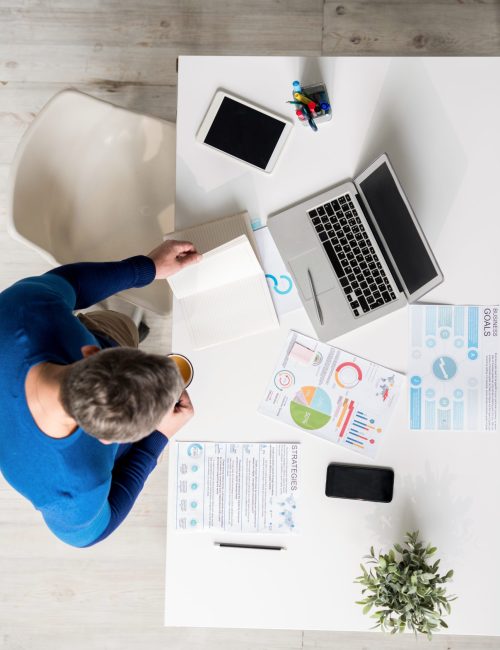 Directly above view of busy young man in blue sweater standing at table and  viewing his notes in diary while drinking coffee and thinking about marketing project in office.